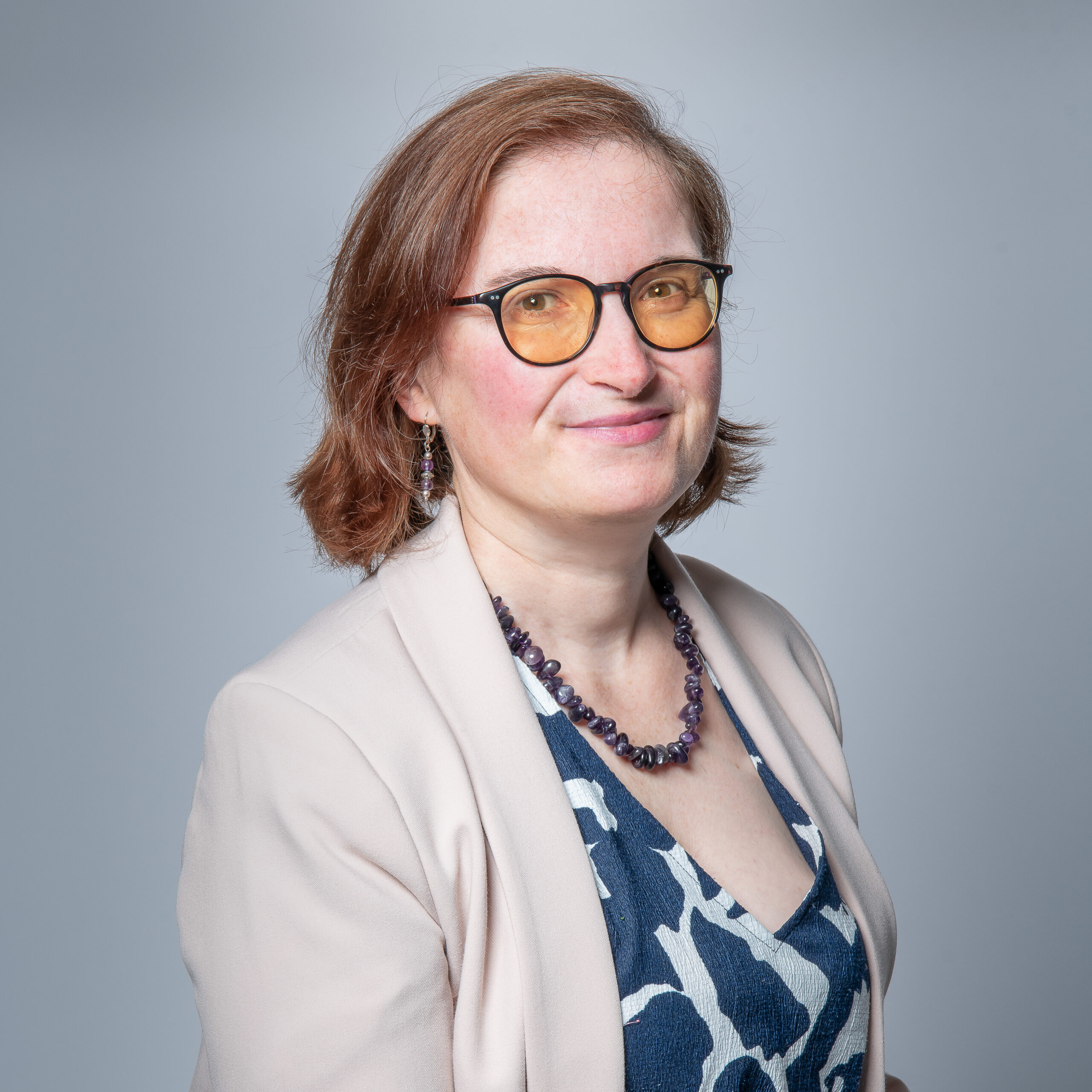 C’est ma photo, un portrait professionnel, j’ai un visage souriant et apaisant. Je porte des lunettes aux verres légèrement teintés, une veste claire sur une robe bleue à motifs blancs, ainsi qu’un collier en perles sombres. Mes cheveux châtains, mi-courts, sont coiffés simplement. Le fond est uni et neutre, mettant en valeur sa posture calme et bienveillante.
